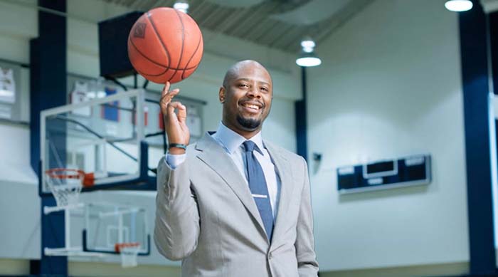 Smiling bald Black man in a gray suit and blue tie stands in a gymnasium, spinning a basketball on his finger