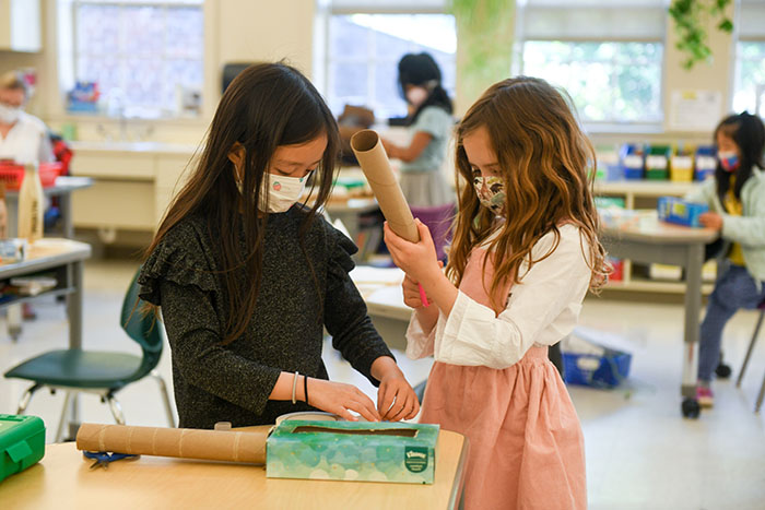 Two Masked Elisabeth Morrow School students working on an invention with cardboard tubes, kleenex boxes and tape in a classroom.