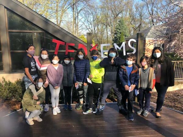 Group of students outside on wooden deck in front of trees and a banner that reads TEDxEMS