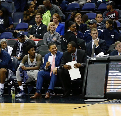 Courtside fans and players at a basketball game, including Patrick Ewing Jr.