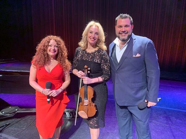 Three musicians on a stage; from left to right a shorter caucasian woman with red curly hair and a red dress, Amelia Gold, a taller blonde woman in a black dress holding a violin, and a tall man with a salt and pepper beard wearing a dark suit
