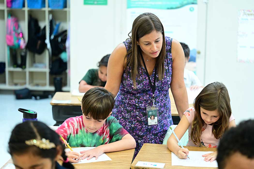 Ms. Samantha Pianko standing behind two students sitting at their desk working.